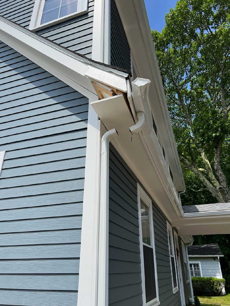 Corner of a two-story house with blue-gray siding where part of the white soffit has broken away and the gutter and downspout are pulled loose